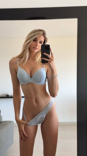 Blonde woman in light blue lingerie takes mirror selfie in bedroom.
