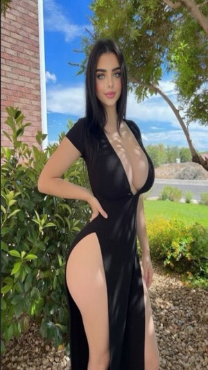 Woman in black dress with high slit posing outdoors near greenery.
