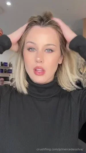 Woman with blonde hair in black turtleneck, hands in hair, indoor setting.
