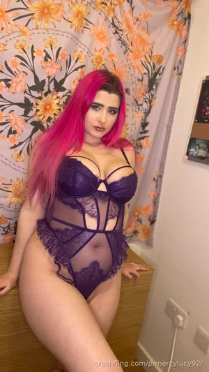 Woman with pink hair in purple lace lingerie posing indoors near floral curtain.