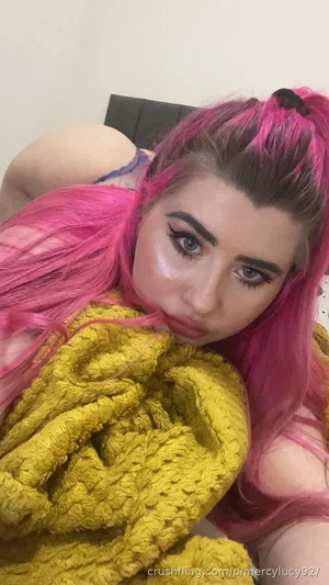 Woman with pink hair, wearing yellow knit top, lying in bed, indoor setting.