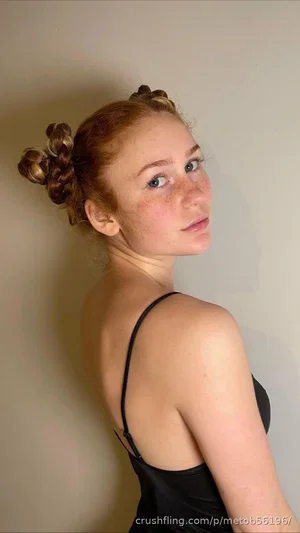 Woman with red hair in braided buns, wearing black spaghetti strap top, posing against plain wall.