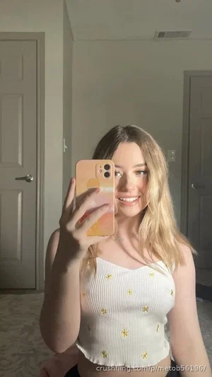 Woman with blonde hair in white crop top taking mirror selfie in bedroom.