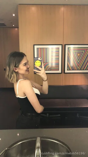 Woman with short blonde hair in black and white top taking mirror selfie in modern bathroom.