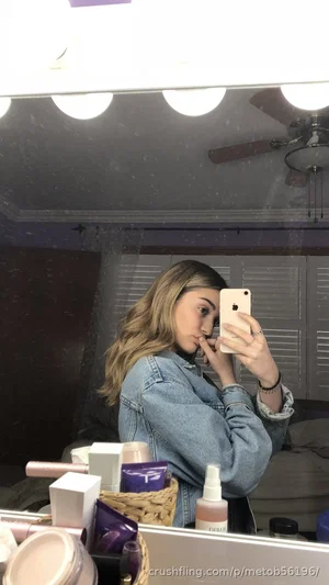 Woman with long hair in denim jacket takes mirror selfie in bedroom with makeup products visible.