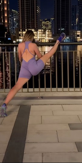 Woman in purple one-piece outfit stretching by waterfront at night, city lights in background.