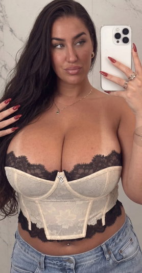 Woman with long dark hair in lace bustier and jeans, taking mirror selfie indoors.