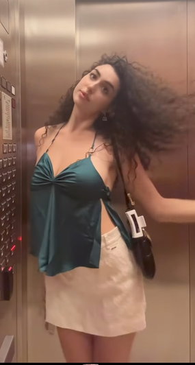 Woman with curly hair in green top and white skirt standing in elevator.