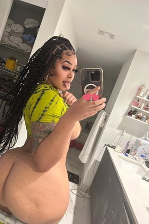 Curvy woman with braids in yellow top, bare buttocks, bathroom mirror selfie.