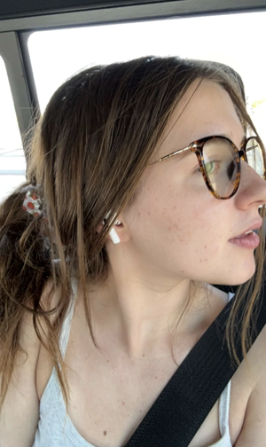Woman with brown hair and glasses in car, wearing white tank top and seatbelt.