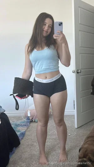 Woman in light blue crop top and black shorts takes mirror selfie in bedroom.