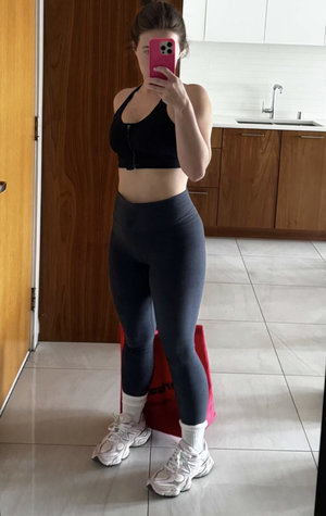 Woman in black sports bra and leggings takes mirror selfie in kitchen.