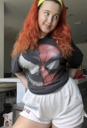 Woman with red hair in a Spider-Man shirt and white shorts posing indoors.
