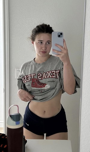 Woman in gray t-shirt and black shorts taking mirror selfie indoors.