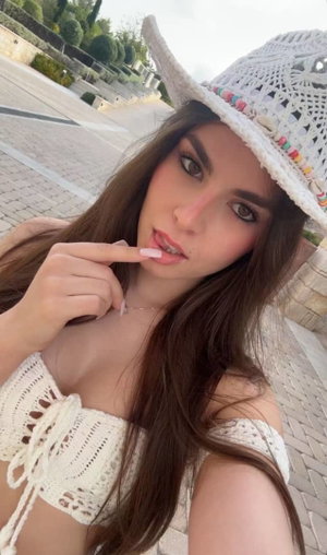 Brunette woman in white crochet top and hat taking outdoor selfie.