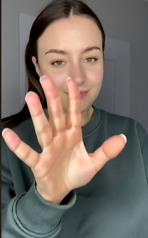 Brunette woman in green sweatshirt waves hand indoors.