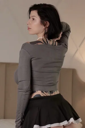 Woman with dark hair and tattoos wears gray top and black skirt, standing in bedroom.
