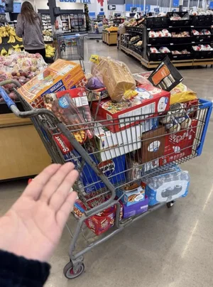 Hand reaching toward a shopping cart filled with groceries in a supermarket aisle.