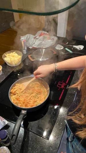Woman with long hair cooking pasta on a stovetop in a kitchen.
