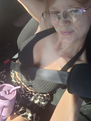 Woman with glasses in black top and patterned shorts sitting in car, holding pink cup.