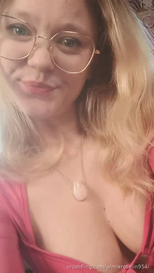 Woman with blonde hair and glasses in a pink top, wearing a pendant necklace, indoors.
