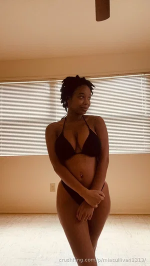 Woman in black bikini standing indoors near window, hands on hips.