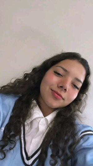 Woman with curly dark hair wearing a light blue jacket and white collared shirt, indoors.