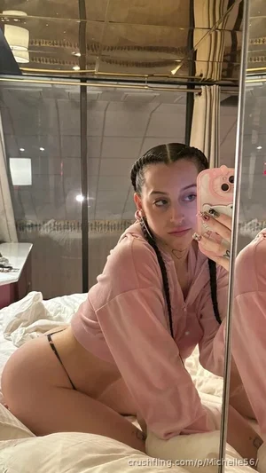 Woman in pink hoodie and black thong posing on bed in mirror selfie.