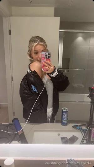 Blonde woman in black jacket and gray top takes mirror selfie in bathroom.