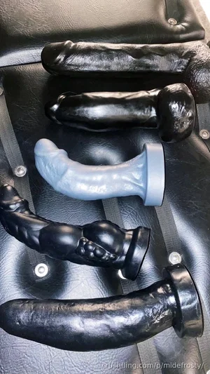 Five black and one light blue dildo toys arranged on a black leather surface with straps.