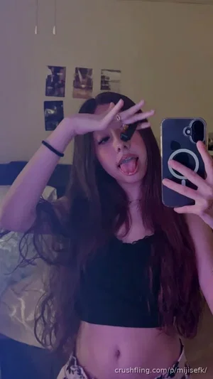 Woman with long dark hair in black crop top and patterned bottoms taking mirror selfie in bedroom.