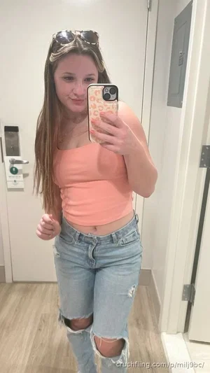 Woman in peach crop top and ripped jeans taking mirror selfie in hallway.