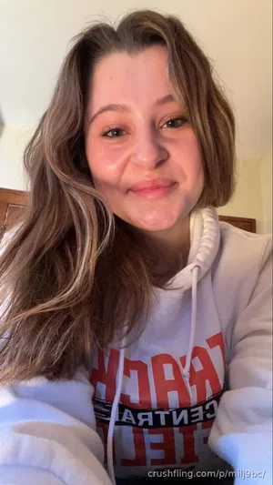 Woman with long brown hair wearing a white hoodie, smiling in a bedroom.