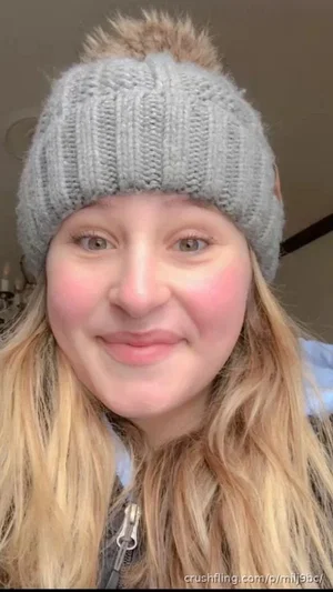 Woman with blonde hair wearing gray knit beanie and jacket, smiling indoors.