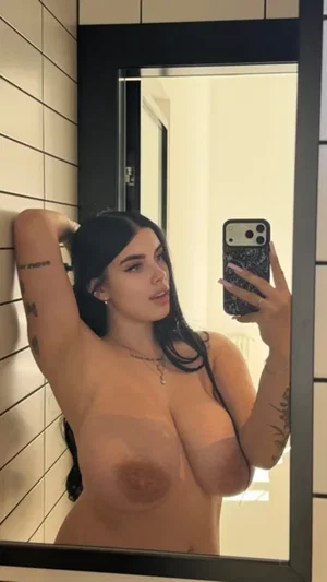 Curvy woman with dark hair taking topless mirror selfie in bathroom.
