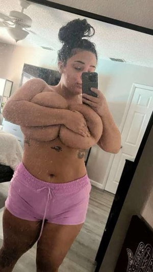 Woman with curly hair in bun, wearing pink shorts, taking mirror selfie in bedroom.