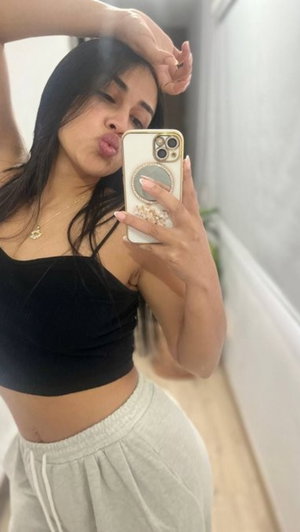 Woman with dark hair in black crop top and gray sweatpants taking mirror selfie indoors.