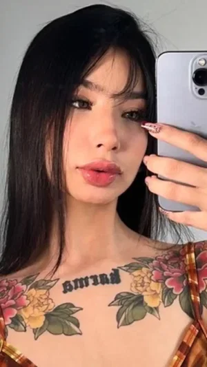 Woman with long black hair and floral chest tattoos taking a selfie.