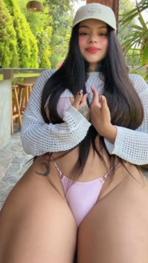 Woman with long dark hair in light pink bikini top and mesh sweater, sitting outdoors.