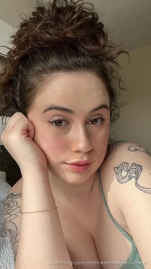 Woman with curly hair and tattoos, wearing a green tank top, resting her head on her hand indoors.
