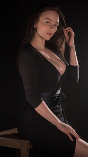 Woman in black dress with deep neckline sitting on wooden stool against dark background.