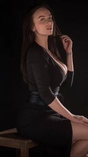 Woman with long dark hair in black dress posing on stool against black background.