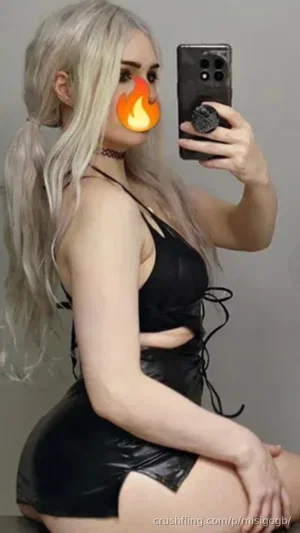 Woman with long blonde hair in black leather outfit taking mirror selfie indoors.