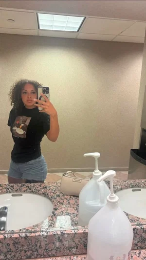 Woman with curly hair in black t-shirt and denim shorts taking mirror selfie in bathroom.