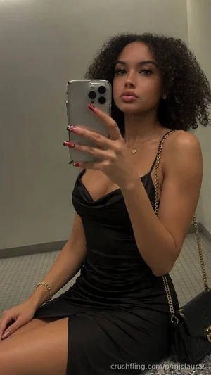 Woman with curly hair in black dress taking mirror selfie, holding phone, sitting on floor.