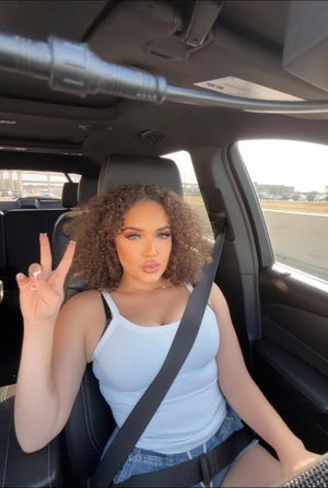 Curvy woman with curly hair in white tank top posing in car.