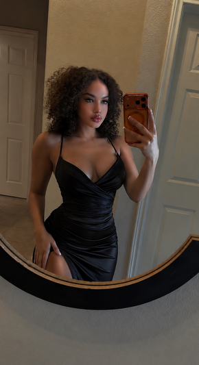 Woman with curly hair in black dress taking mirror selfie indoors.