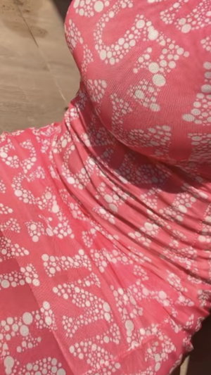 Woman in pink patterned dress indoors on wooden floor.