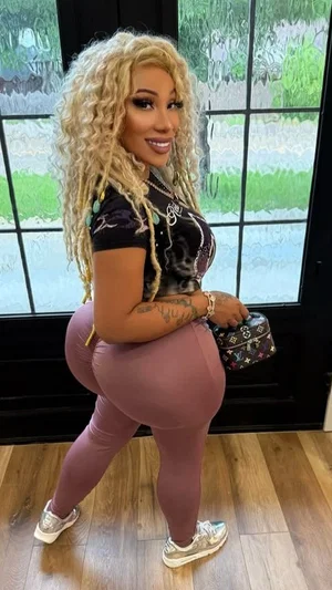 Woman with curly blonde hair in tight pink leggings and black top, standing indoors near glass door.