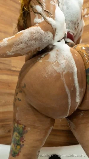 Woman with tattoos covered in soap suds, showing back and buttocks, in bathroom setting.